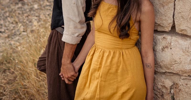 ידיים שלובות ליד קיר אבן, שמלה צהובה — פרט מצילומי זוגיות
