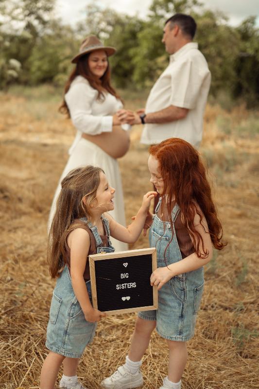 שתי ילדות עם שלט BIG SISTERS וההורים מאחור – צילומי הריון משפחתיים